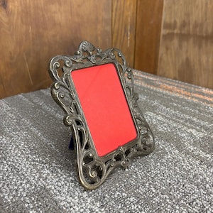 May include: Ornate, antique-style picture frame with a silver finish. The frame features intricate scrollwork and a rectangular opening with a red backing. The frame is designed to stand upright and is suitable for displaying a photograph or artwork.