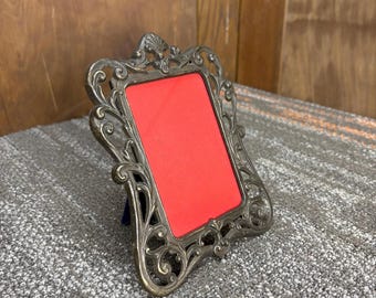 Può includere: Cornice per foto in stile antico con finitura argentata e dettagli ornati. La cornice presenta un'apertura rettangolare con un retro rosso. La cornice è progettata per stare in piedi ed è adatta per esporre una fotografia o un'opera d'arte.