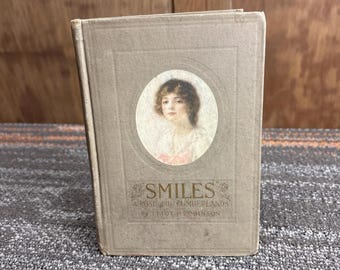 Puede incluir: Un libro antiguo titulado "Smiles" de Leigh H. Robinson. El libro tiene una cubierta gris claro con un retrato de una mujer en un marco ovalado. El título está en escritura decorativa con acentos florales. El libro está ligeramente inclinado.