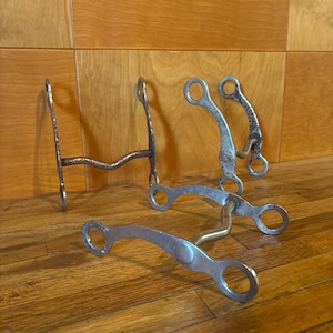 May include: Three metal horse bits with curved mouthpieces and side shanks. Each bit features circular rings at the ends of the shanks for attachment. The metal surfaces show signs of wear and texture, with a mix of silver and brass tones.