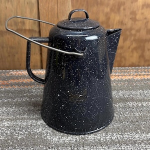 Puede incluir: Una cafetera de esmalte negro con motas blancas. La cafetera tiene una tapa con un asa pequeña y un asa de metal que se arquea sobre la parte superior. La cafetera está sobre una superficie gris y naranja estampada.