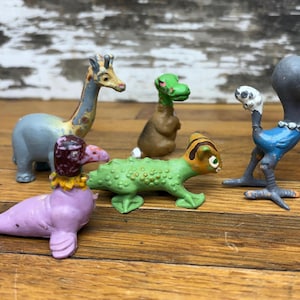 May include: A collection of colorful, vintage toy figurines. The set includes a giraffe, a seal-like creature, a dinosaur, a creature with an elephant-like head, and a small dinosaur. The toys are painted in various colors, including blue, green, purple, and brown.