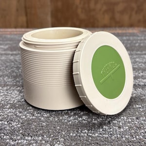 May include: A beige Thermos food container with a matching lid. The lid has a green center with the Thermos logo in white. The container is cylindrical with a ribbed exterior. The lid is off to the side.