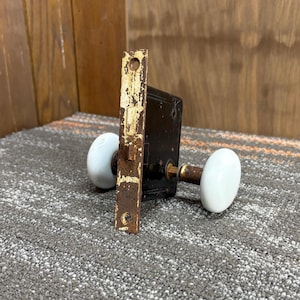 以下が含まれることがあります： 黒い金属製の箱、錆びた金属板、2つの白い楕円形の磁器製ドアノブを備えた、アンティークのドアロックセット。 金具には、塗料の剥がれや錆など、経年劣化の兆候が見られます。