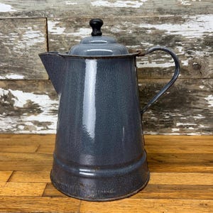 Puede incluir: Una cafetera de esmalte gris con un pomo negro en la tapa y un asa de metal. La cafetera está sobre una superficie de madera.