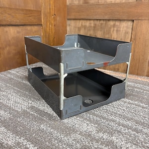 May include: A two-tiered, gray metal desk organizer. The trays are rectangular with rounded corners and a cutout on the front. The organizer is supported by silver metal legs. The background is a wooden wall and a gray carpet.