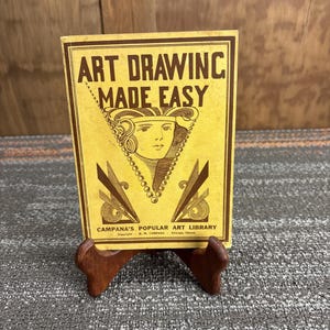 May include: A vintage art instruction book titled "ART DRAWING MADE EASY" on a wooden display stand. The book has a yellow cover with brown text and illustrations. The book is from Campana's Popular Art Library, Chicago, Illinois.