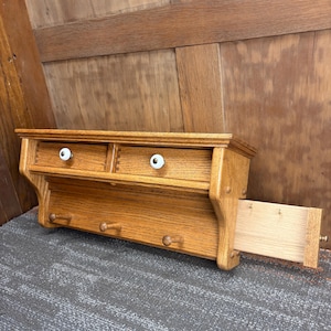 May include: A wooden wall-mounted shelf with two small drawers and white knobs. Below the drawers are three wooden pegs for hanging items. The shelf is made of light brown wood and has a drawer pulled out.