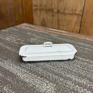 Puede incluir: Un recipiente rectangular de metal blanco con tapa y asa. El recipiente tiene un aspecto ligeramente desgastado con zonas de color óxido en los bordes. El fondo es una superficie de madera y una alfombra gris.