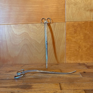 May include: Two silver-colored metal surgical instruments with ring handles. One instrument is positioned vertically and features straight, narrow blades. The second instrument is positioned horizontally and features curved, narrow blades.