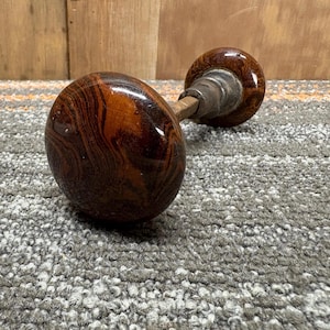 May include: A pair of antique door knobs with a swirled brown and tan pattern. The knobs are made of a glossy material and are connected by a metal rod. The background is a wooden surface and a gray carpet.