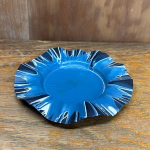 May include: A small, round, blue dish with a ruffled edge. The dish has a dark blue and white pattern around the rim. The center of the dish is a solid, lighter blue color. The dish is sitting on a wooden surface.