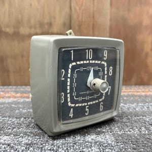 Puede incluir: Un reloj General Electric vintage con una caja cuadrada gris. La esfera del reloj es negra con números blancos y una esfera blanca. Tiene una perilla en la parte delantera y una pequeña pieza de plástico en la parte superior.