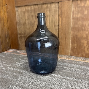 May include: A large, dark blue glass bottle with a rounded body and a long neck. The bottle is made of thick glass and has a slight gradient in color, with a darker shade at the base. It is likely used for decorative purposes.