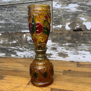 May include: A small amber glass oil lamp with a decorative floral design in red, green, and yellow. The lamp has a round base and a tall, slender neck.