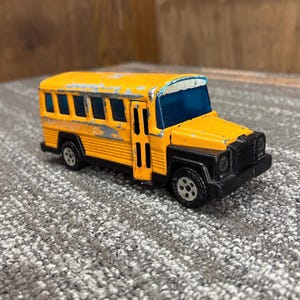 May include: A vintage yellow toy school bus with black accents. The bus has visible wear and tear, including paint chipping. The windows are blue, and the wheels are black. The front of the bus has a black grill and headlights.