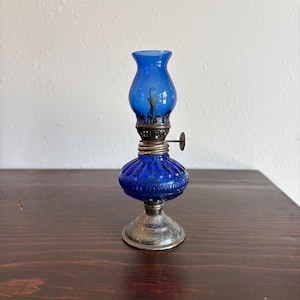 May include: A small, vintage oil lamp with a vibrant cobalt blue glass shade and base. The lamp features a silver-colored metal base and intricate detailing around the wick holder. The lamp is a decorative item.