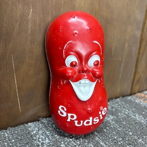 Puede incluir: Una figura roja sonriente de dibujos animados con la palabra "Spudsle" en escritura blanca. La figura tiene ojos y dientes blancos y tiene forma de patata. La figura tiene pequeños agujeros en su superficie.