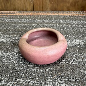 May include: A small, round, pink ceramic bowl with a smooth, matte finish and a thick, curved rim. The bowl has a deep, concave center and a uniform, solid pink color throughout its exterior and interior surfaces, presenting a simple, circular form.
