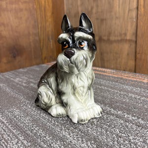 May include: A ceramic Schnauzer figurine in a seated position. The dog has a black and gray coat with a white chest and paws. It features detailed facial features, including brown eyes and a dark nose. The figurine is on a gray surface.