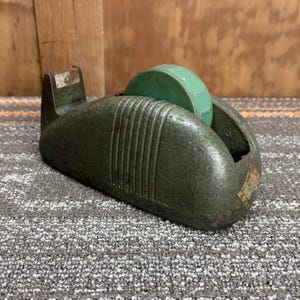 Puede incluir: Dispensador de cinta adhesiva verde vintage con un rollo de cinta verde. El dispensador tiene un diseño industrial aerodinámico con ranuras verticales y una pequeña etiqueta. El dispensador es de metal y muestra signos de desgaste.
