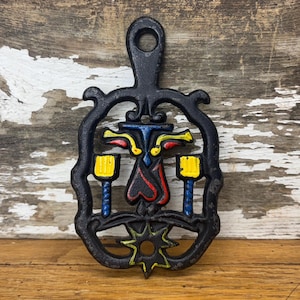 Puede incluir: Un salvamanteles de hierro fundido negro con un diseño amarillo, rojo y azul. El salvamanteles tiene una forma de estrella en la parte inferior y un asa de bucle en la parte superior.