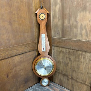 May include: A vintage wooden weather station with a thermometer, barometer, and small clock. The barometer has a silver face with gold trim and a needle pointing to "Fair". The thermometer is marked in Fahrenheit.