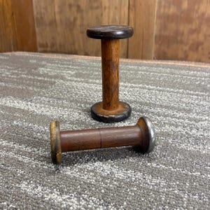 Puede incluir: Dos carretes de madera antiguos. Un carrete está de pie, mientras que el otro está en posición horizontal. Los carretes son de madera marrón oscuro con forma cilíndrica. El carrete vertical mide unos 7,6 cm de alto.