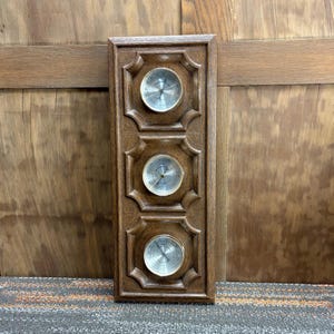 Puede incluir: Una estación meteorológica de madera vintage con tres medidores circulares. Los medidores tienen caras plateadas y bordes dorados. El marco de madera es de color marrón oscuro y tiene un diseño ornamentado.