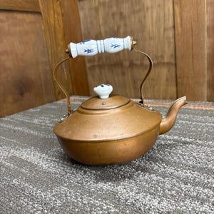 May include: A copper tea kettle with a white and blue porcelain handle and lid knob. The kettle has a curved spout and brass-colored metal supports. The handle is decorated with blue floral designs.