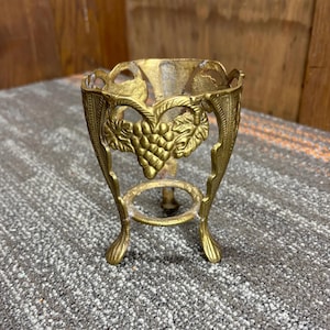 May include: A vintage brass egg cup with a decorative grape and leaf design. The egg cup has a round base and three curved legs. The top has an openwork design with a grape cluster in the center.