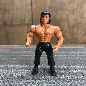 May include: A vintage action figure of a muscular man with black hair and a red headband. The figure is wearing black pants and boots. The figure has a tan skin tone and is posed in a standing position.