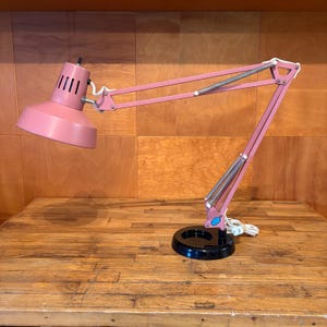 May include: A pink desk lamp with an adjustable articulated arm and a wide, conical shade. The lamp features a round black base, exposed metal springs for tension, and a white power cord. The shade has vertical ventilation slits near the top.