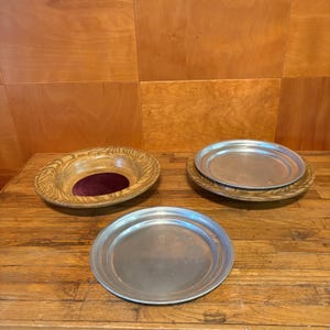 May include: A wooden bowl with a dark purple velvet center insert, alongside two round silver-colored metal plates. One plate rests inside a matching wooden bowl, while the other sits independently on the surface in the foreground.