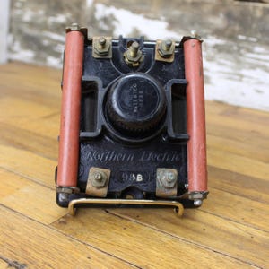 Puede incluir: Un componente eléctrico negro con el texto "Northern Electric" y "Patented 1938" en él. El componente tiene dos aisladores rojos a cada lado. El componente tiene el número "93B" en él.