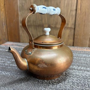 May include: A copper tea kettle with a white and blue handle and a white knob on the lid. The kettle has a spout and a rounded body. The handle is arched and attached to the body with copper brackets. The kettle is sitting on a gray surface.