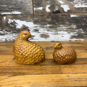 May include: Two ceramic quail figurines, one larger than the other. The larger quail is brown with a red crest and the smaller quail is brown. Both quail are sitting on a wooden surface.