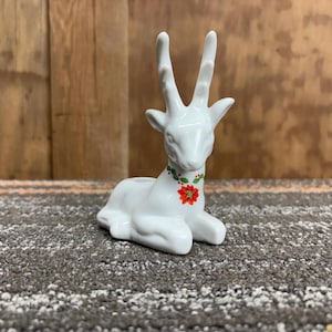 Puede incluir: Figura de reno de cerámica blanca con cuernos altos y una flor de pascua roja en el pecho. El reno está en posición sentada, con un collar floral verde y rojo. La figura es un artículo decorativo.