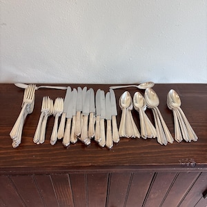 May include: A large collection of silver-toned flatware including multiple forks, knives, and spoons arranged in organized groups. The utensils feature a simple, smooth handle design with a polished metallic finish, reflecting light across the set.