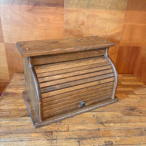 May include: A rectangular wooden bread box with a medium brown finish and a visible wood grain. The container features a curved, slatted roll-top door that slides upward, equipped with a small, round wooden knob for opening and closing the unit.