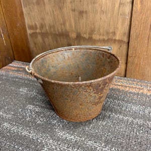 May include: A small, rusty metal bucket with a wire handle. The bucket has a tapered shape and is heavily weathered, showing signs of rust and age. The background includes a wooden surface and a gray and orange patterned rug.