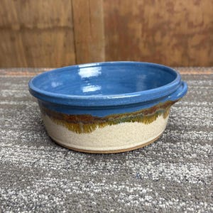 May include: A handmade ceramic bowl with a blue interior and rim, and a tan exterior. The rim has a brown and green band. The bowl has a round shape and two small handles. The bowl is suitable for serving food.