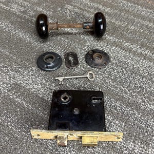 May include: An assortment of antique door hardware, including a black doorknob, escutcheons, a key, and a mortise lock. The hardware shows signs of age and wear, with a vintage aesthetic. The components are arranged on a textured surface.