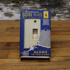 以下が含まれることがあります： 青と白の箱に入ったヴィンテージのライトスイッチプレート。箱には「Luminous Guide Plate - Glows in the Dark」と書いてあり、灯台の絵が描かれています。スイッチプレートは白で、スイッチが1つ付いています。