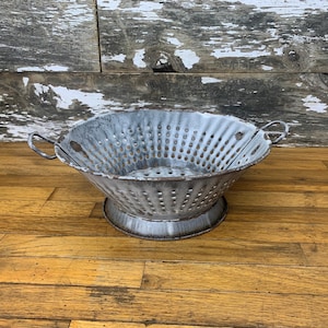 Puede incluir: Un colador de metal envejecido con un acabado gris y blanco. El colador tiene forma de cuenco ancho y poco profundo con numerosos agujeros pequeños y dos asas curvas. Se asienta sobre una base baja y circular, ideal para la cocina.