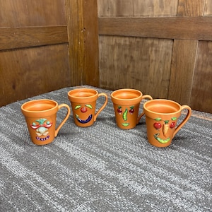 Peut inclure: Quatre tasses en céramique orange avec des anses, chacune décorée d'un visage de légume unique. Les visages comprennent des tomates, des carottes et des pois. Les tasses sont disposées sur une surface grise.