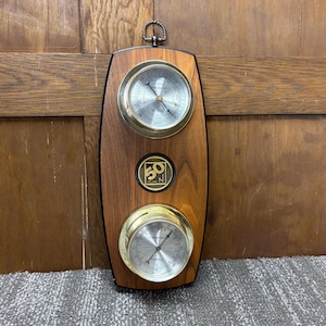 May include: A vintage wooden weather station with two brass-rimmed gauges and a circular emblem. The gauges display pressure and temperature readings. The wooden frame has a dark finish and a metal loop for hanging.