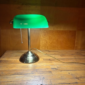 May include: A banker's lamp featuring a polished brass base and a vibrant green glass shade. The lamp includes a vertical stand, a pull chain switch, and a thin electrical cord extending from the base to provide power for the light.