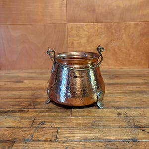 May include: A small, round, hammered copper pot featuring a metallic finish with a warm, reddish-brown hue. The vessel includes a curved metal handle attached to two side loops and rests on three small, sturdy metal feet at the base.