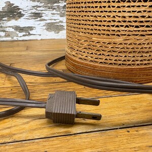 May include: A cylindrical lampshade made from corrugated cardboard sits on a wooden surface. A brown electrical cord and plug are in the foreground. The lampshade has a natural, textured appearance.